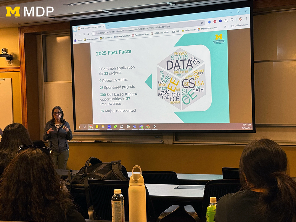 Female program manager standing in front of a projection screen with project information displayed behind her, being observed by students in auditorium-style seating. 