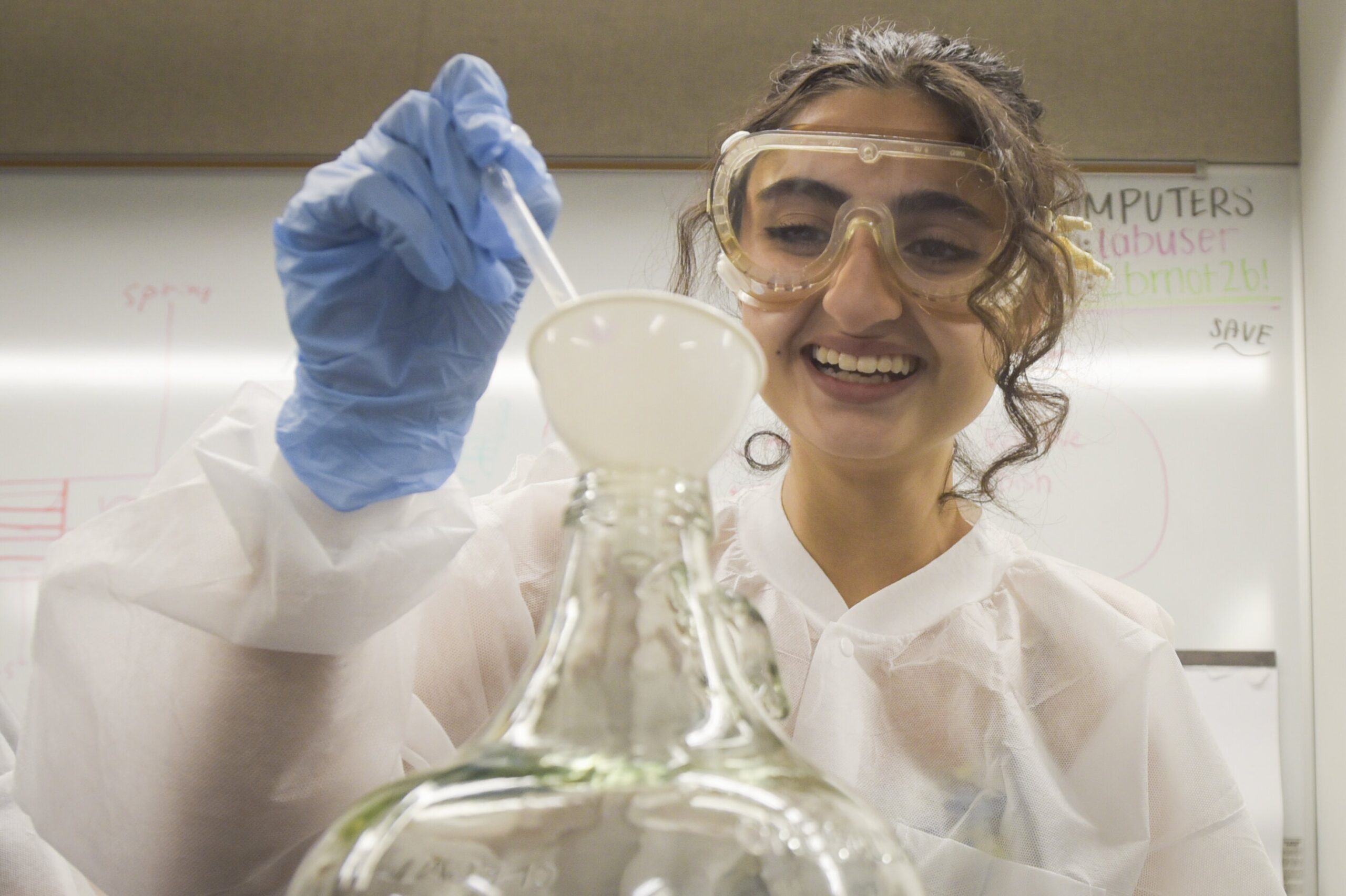 Student in lab attire smiling while pipetting liquid into glass jar