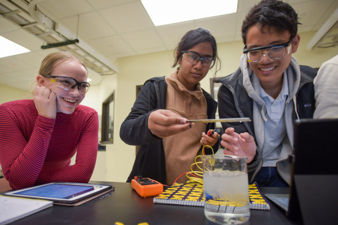 Three students wearing goggles looking over beaker with ice and wires during lab
