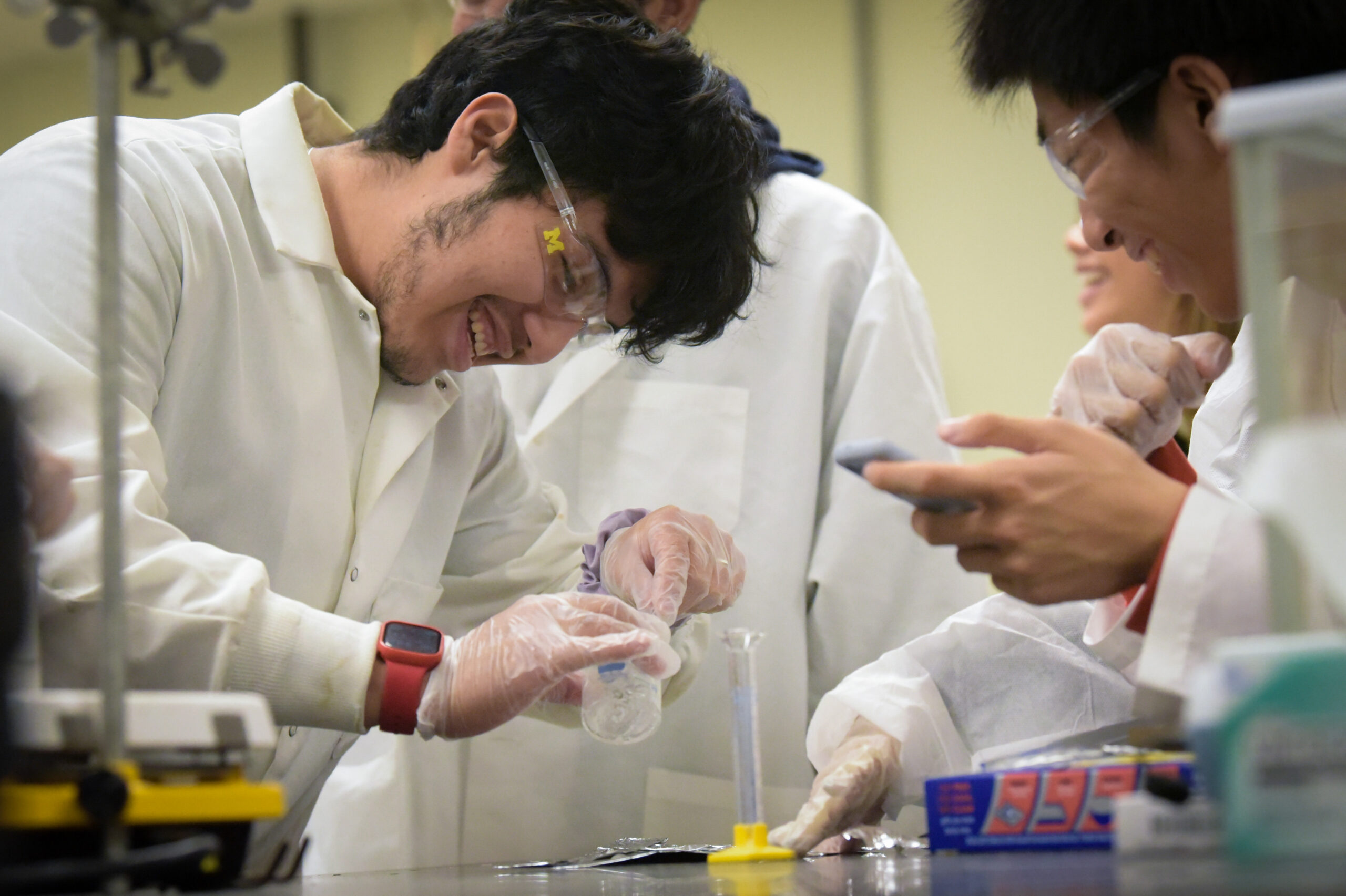 Students in lab attire smiling while looking at contents of beaker