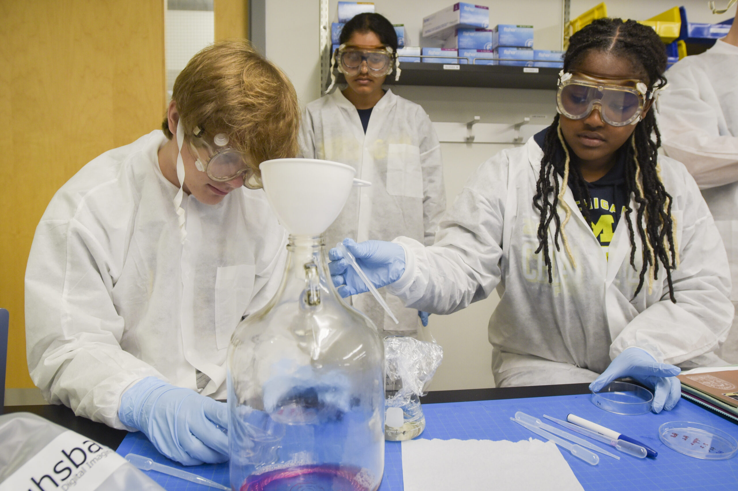 Students in lab attire side by side removing liquid from petri dish