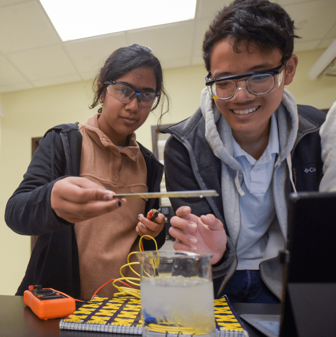 Two students in safety goggles looking over beaker conducting experiment